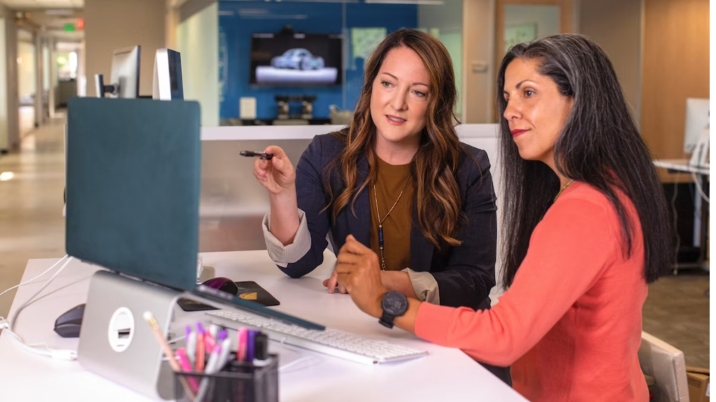 two professional looking women, sit at a computer. One woman with a navy blazer instructs the other in a coral blazer
