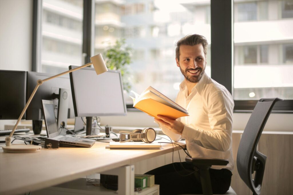a man in business attire at an office computer desk, holding a book and looking optimistic