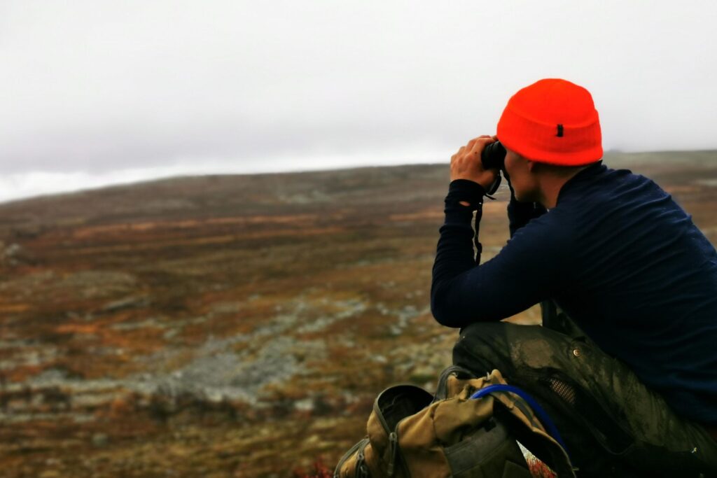 a man in camouflage gear, looks out at a grassland scene via binoculars